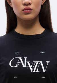 Μαύρο βαμβακερό T-shirt με μεγάλο λευκό γραφικό κείμενο που αναγράφει έντονα "CALVIN" και μικρότερα γράμματα "NEW," "KLEIN," και "YORK" από κάτω.