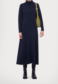 Navyblaues Langarmkleid mit hohem Rollkragen, aus weichem Stoff. Kombiniert mit einer grünen Schultertasche und glänzenden schwarzen Knöchelstiefeln.