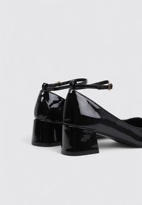 Chaussures à talon bloc en cuir verni noir avec une bride au niveau de la cheville, présentant une finition lisse et brillante ainsi qu'une forme de bout triangulaire.
