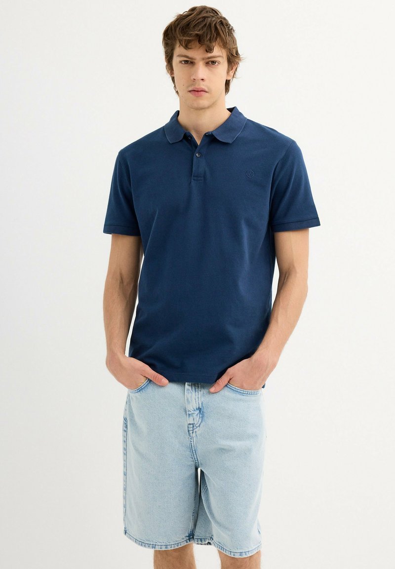 Polo de algodón azul marino con cuello, mangas cortas y un logo sutil; combinado con pantalones cortos de mezclilla azul claro.