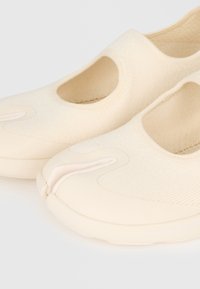 Par de sapatos slip-on bege com design de dedo dividido e material superior em malha, mostrados contra um fundo claro e simples.