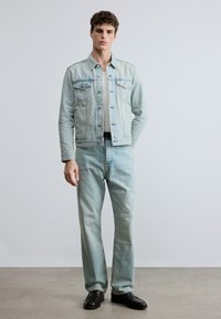 Ljusblå jeansjacka med två bröstfickor, kombinerad med matchande ljusblå jeans och svarta läderskor, stående mot en neutral bakgrund.