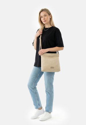 Naine musta t-särgi, helesiniste teksade ja valgete tossude ning reguleeritava rihmaga beežist ristihoidjaga koti taustal valge taust.