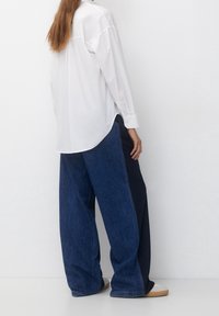 White oversized button-up shirt made of cotton, paired with wide-leg dark blue denim pants. Accessories include beige slip-on shoes.
