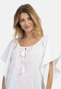 Hvid bomuldsbluse med korte flæser på ærmerne, prydet med pink blomsterbroderi og en frynseknytte ved halsudskæringen.