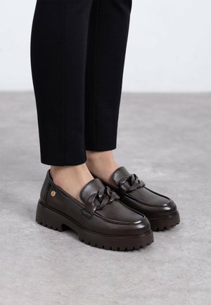 Mocasines de cuero marrón con suela gruesa, con un acento trenzado en la parte frontal. Superficie texturizada y detalle del logo dorado en el lateral.