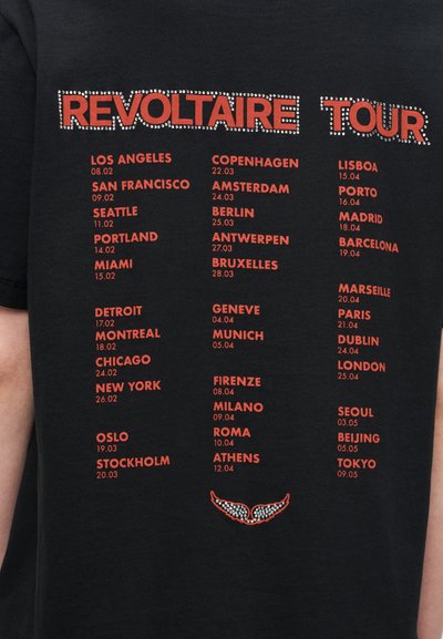 Μαύρο μπλουζάκι με κόκκινο κείμενο που αναγράφει πόλεις και ημερομηνίες για την "REVOLTAIRE TOUR" με διακοσμητικά από στρας στο κάτω μέρος.