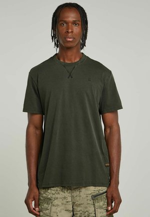 Camiseta de manga corta verde oliva hecha de algodón con cuello redondo, que presenta un detalle en forma de X cosido y un pequeño logo en el pecho.