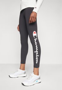 Leggings grigi con il logo bianco "Champion" stampato di lato. Indossati con scarpe da ginnastica bianche con accenti blu e texture in rete.