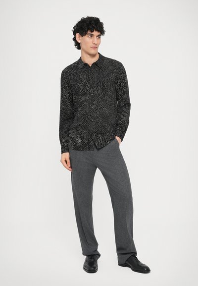 Schwarzes Hemd mit Knöpfen und Polka-Dot-Muster, kombiniert mit grauer Hose, vor neutralem Hintergrund präsentiert.