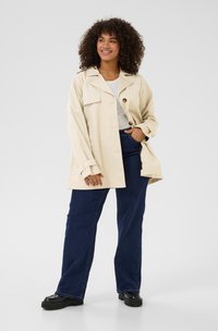 Beige oversized jack met knoopsluiting, schouderflap en lange mouwen, gecombineerd met donkerblauwe straight-leg jeans en zwarte plateau schoenen.