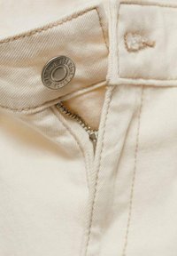 Kremasti pamučni traper s metalnim zatvaranjem na dugme i zatvaračem. Ima vidljive šavove i malu oznaku od tkanine. Glatka tekstura.