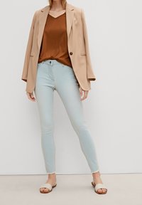 Beiger Blazer über einem braunen V-Ausschnitt-Oberteil, kombiniert mit hellblauen Skinny-Jeans und hautfarbenen Sandalen mit gedrehtem Stoffdetail.