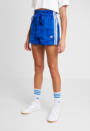 Osoba ubrana w niebieskie spodenki Adidas z białymi paskami, białe skarpetki Adidas z niebieskimi paskami, białe buty sportowe oraz krótką białą bluzkę.