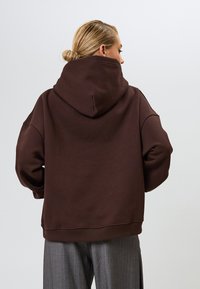 Barna, túlméretezett kapucnis pulóver nagy kapucnival, széles ujjal és bordázott aljjal, szürke csíkos nadrággal párosítva. Nincsenek látható minták vagy fémszerelvények.