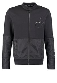 Schwarze Bomberjacke mit einem Reißverschluss vorne, hohem Kragen und einer Mischung aus Stofftexturen. Verfügt über seitliche Reißverschlusstaschen und ein dezentes Logo auf der Brust.