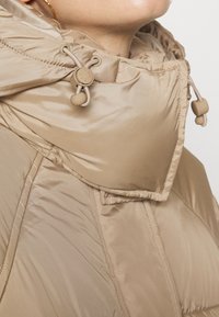 Beige polstret jakke med høj krave, justerbare snore og glat, skinnende stof. Har syningsmønstre og en afslappet pasform.