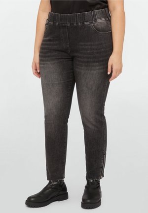 Vrouw draagt vervaagde zwarte slim-fit jeans met elastische tailleband en ritsen bij de enkels, gecombineerd met zwarte enkellaarsjes en een zwarte top.