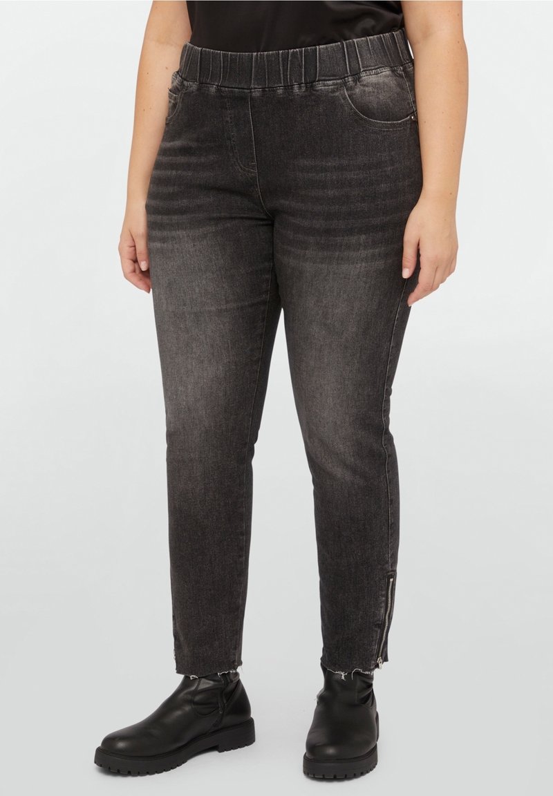 Femme portant un jean slim noir délavé avec une taille élastique et des fermetures éclair aux chevilles, associé à des bottines noires et un haut noir.