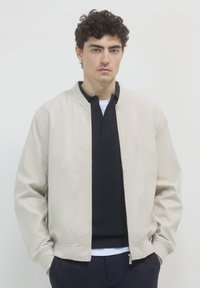 Giovane con i capelli ricci che indossa una giacca beige con zip sopra un maglione scuro a costine e pantaloni blu, in piedi davanti a uno sfondo chiaro e uniforme.