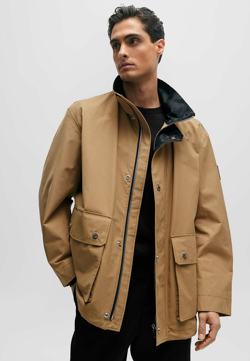 Braune wasserdichte Jacke mit hohem Kragen, Reißverschluss vorne, schwarzer Innenauskleidung und zwei großen Vorder-Taschen mit Druckknopfverschlüssen.