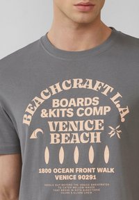 Grijze katoenen t-shirt met vervaagde roze grafische tekst, ronde hals en korte mouwen, met strandthema-elementen.