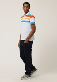Polo shirt in het wit met horizontale strepen in rood, geel, blauw en turquoise; gecombineerd met donkerblauwe jeans en sneakers met grijze en groene accenten.