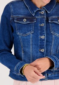 Vrouw in een blauwe denimjack die dichtgeknoopt is, met gouden kralenarmbanden om haar linkerstuk, met de handen voorzichtig voor zich samengevouwen.