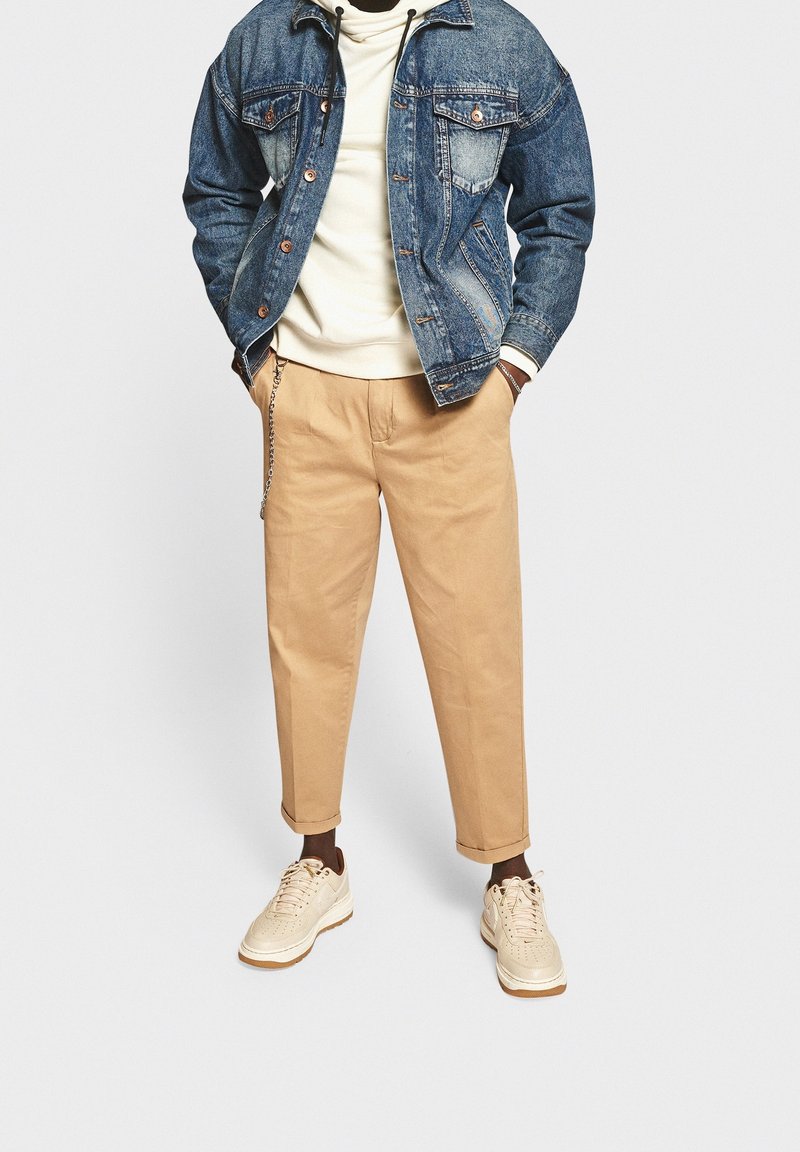 Giubbotto di jeans con una finitura blu sbiadita, felpa bianca sotto, pantaloni beige aderenti e sneakers beige con dettagli testurizzati.