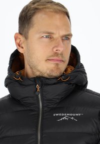 Svart puffjacka med hög krage, med en brun insida, dragkedjestängning och "SWEDEMOUNT" logotyp broderad på bröstet.