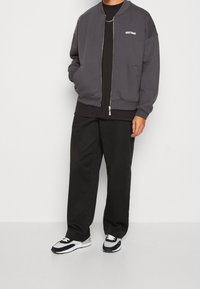 Graue Bomberjacke mit Reißverschluss, gerippten Bündchen und Seitentaschen, getragen über einem schwarzen Hemd, kombiniert mit schwarzen lässigen Hosen und Sneakers.