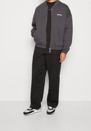 Graue Bomberjacke mit Reißverschluss, gerippten Bündchen und Seitentaschen, getragen über einem schwarzen Hemd, kombiniert mit schwarzen lässigen Hosen und Sneakers.