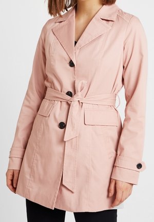 Femme portant un trench-coat rose clair ceinturé avec des boutons noirs et des poches à rabats, debout devant un fond blanc uni.