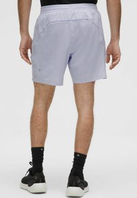 Man in lichtpaarse sport shorts, zwarte sokken en zwarte sneakers, gezien van achteren tegen een effen witte achtergrond.