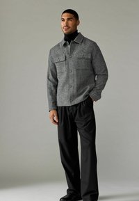 Cămașă gri texturată cu guler, având două buzunare pe piept, purtată cu pantaloni negri largi și pantofi negri, stând pe un fundal neutru.