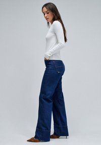 Haut blanc à manches longues associé à un jean large taille haute bleu foncé avec un subtil accent doré sur les boutons et un ourlet droit.