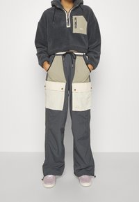 Fleecekort hoodie i mörkgrått med dragkedja och bröstficka. Löst sittande cargobyxor i grått med beige detaljer och flera fickor.