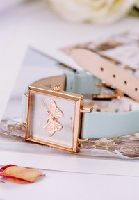 Montre rectangulaire dorée avec un motif de papillon sur un bracelet en cuir bleu clair, placée sur un fond floral.