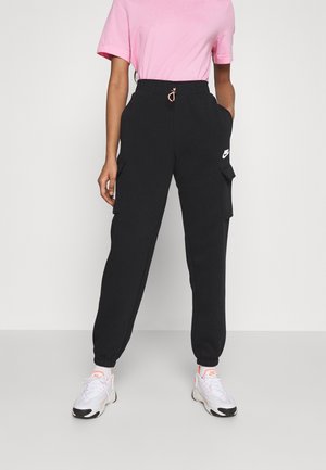 Osoba ubrana w czarne spodnie dresowe Nike cargo, białe i różowe buty Nike oraz jasnoróżową koszulkę z krótkim rękawem na jednolitym tle.