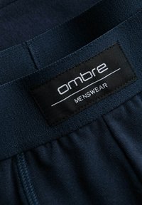 Ceinture bleu foncé avec une texture élastique et une étiquette noire. L'étiquette indique "ombre MENSWEAR" en lettres blanches, sur un fond de tissu lisse.