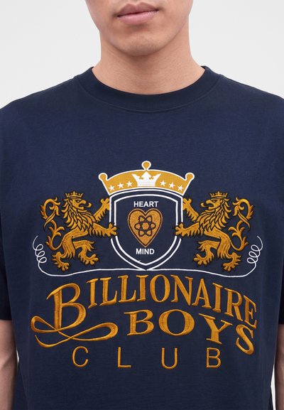 Homme portant un t-shirt bleu marine avec des lions brodés en or, un écusson couronné et le texte "Heart Mind" et "Billionaire Boys Club."