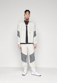 Track suit made of lightweight fabric; cream jacket with gray accents, full zip, and elastic cuffs; matching pants with tapered legs.