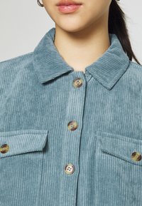 Primo piano di una persona che indossa una camicia di velluto a coste color teal con grandi bottoni marroni e due tasche sul petto con bottone.