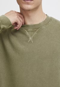 Olive-green sweatshirt with a ribbed neckline and X-stitch detail at the collar. Soft, textured fabric with long sleeves.