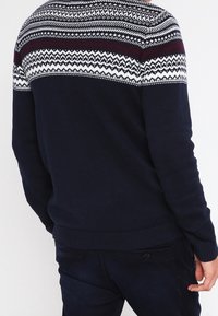 Marinblå stickad tröja med ett mönstrat yoke med vita och vinröda mönster. Ribbad mudd och nederkant ger en texturerad finish.