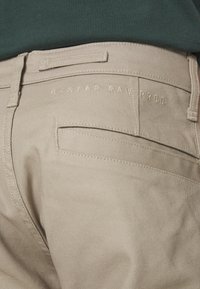 Poche arrière de pantalon beige avec un texte brodé subtil « G-STAR RAW PROD » près de la ceinture, portée avec un haut vert foncé.