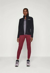 Zwarte fleece jas met rits en logo, grijs shirt, bordeauxrode leggings en grijze sneakers met zwarte accenten. Staande positie tegen een effen achtergrond.