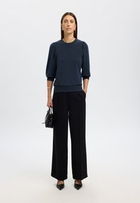 Pullover blu navy di alta qualità con maniche a sbuffo; abbinato a pantaloni neri a gamba larga e décolleté neri a punta. Dettaglio della borsa a tracolla.