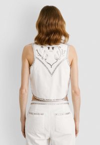 Bijela bezrukavna majica s srebrnim ukrasima i složenim perlicama na leđima, koja ima geometrijski dizajn i blago skraćeni oblik.