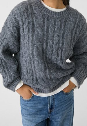 Persoon draagt een dikke grijze kabelgebreide trui over een wit topje, gecombineerd met blauwe jeans en handen nonchalant in de zakken.